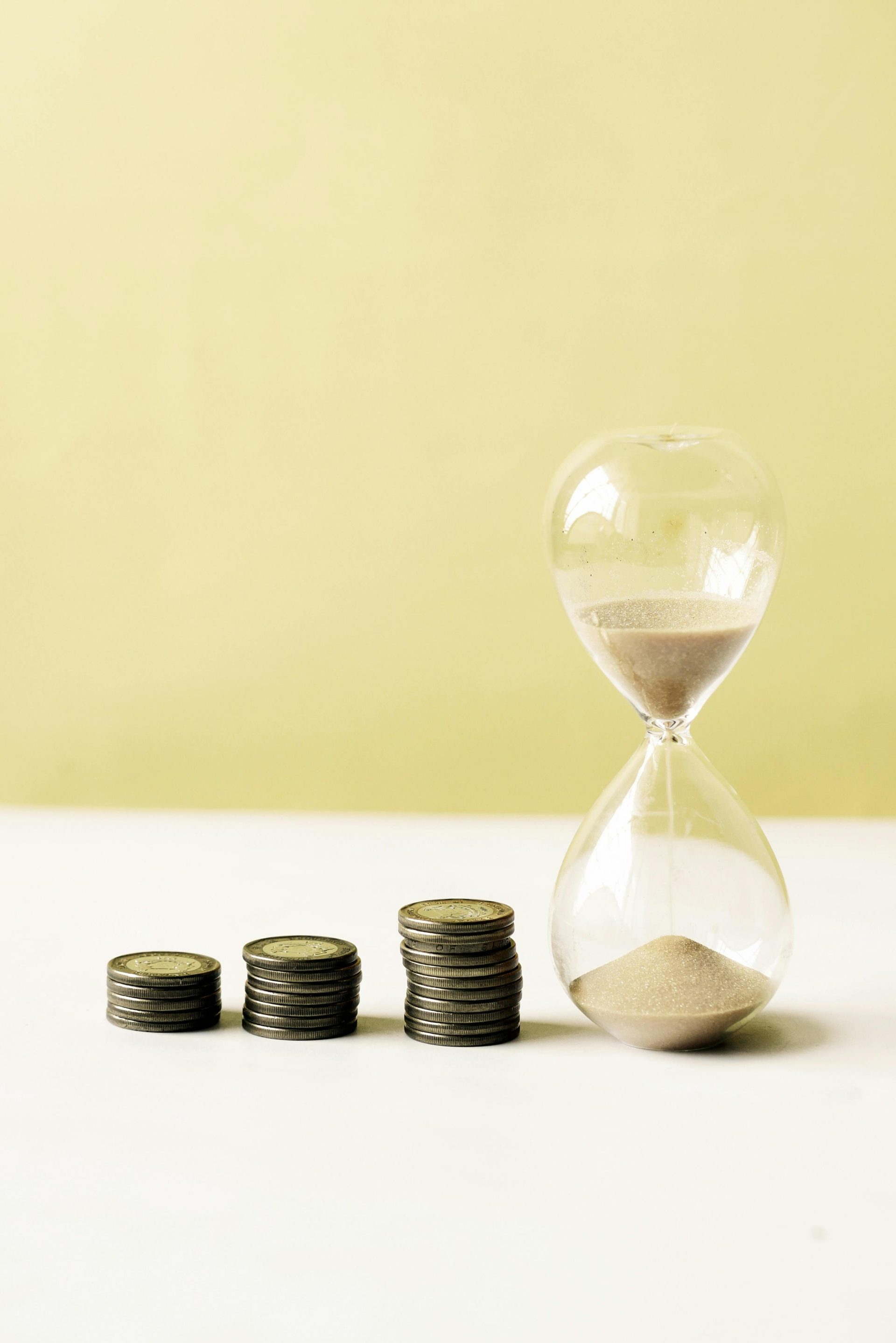 Eine vertikale Nahaufnahme von Münzen und einer Sanduhr vor gelbem Hintergrund, die Zeit und Geld symbolisieren.